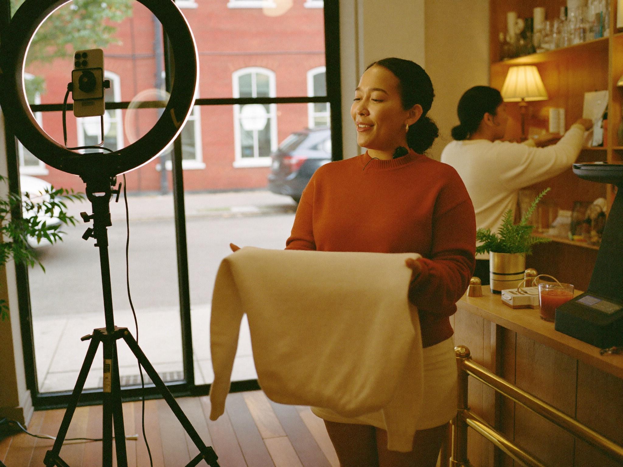 A live selling host mid-sentence to a phone on a tripod, holding a garment up to the camera, with a shop floor and a second person behind her.