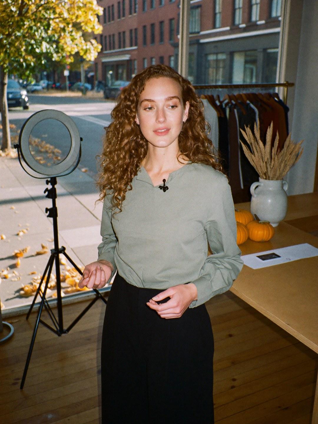 A live selling host mid-conversation in a boutique with small pumpkins and dried wheat on the counter and fallen leaves outside.
